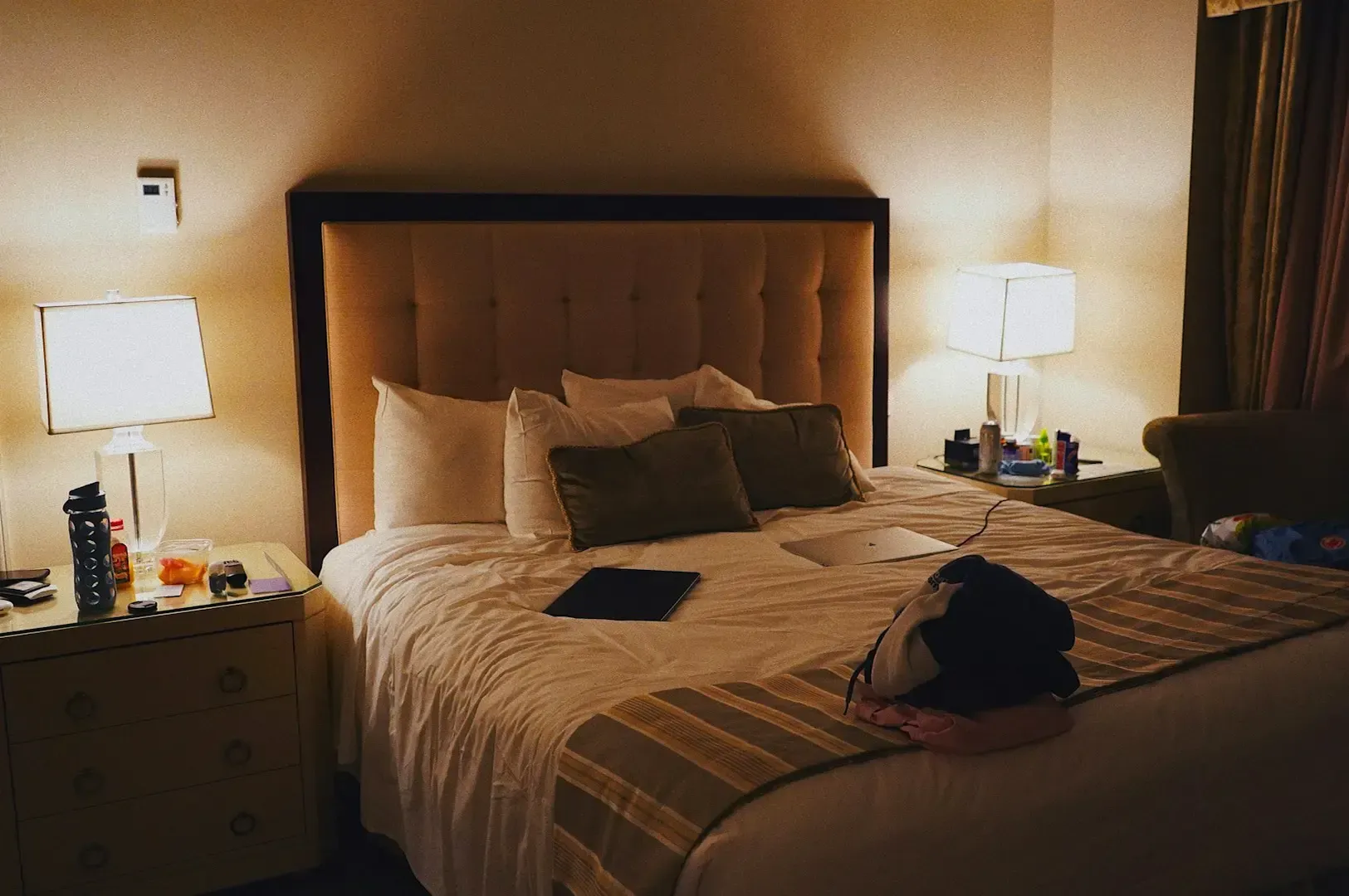 A dimly lit hotel room with a slightly ruffled up bed and two lamps on either side.