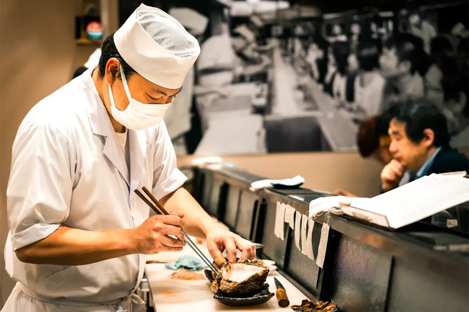 A Japanese chef preparing a seafood dish with chopsticks beside a countertop with a few customers dining in the background.