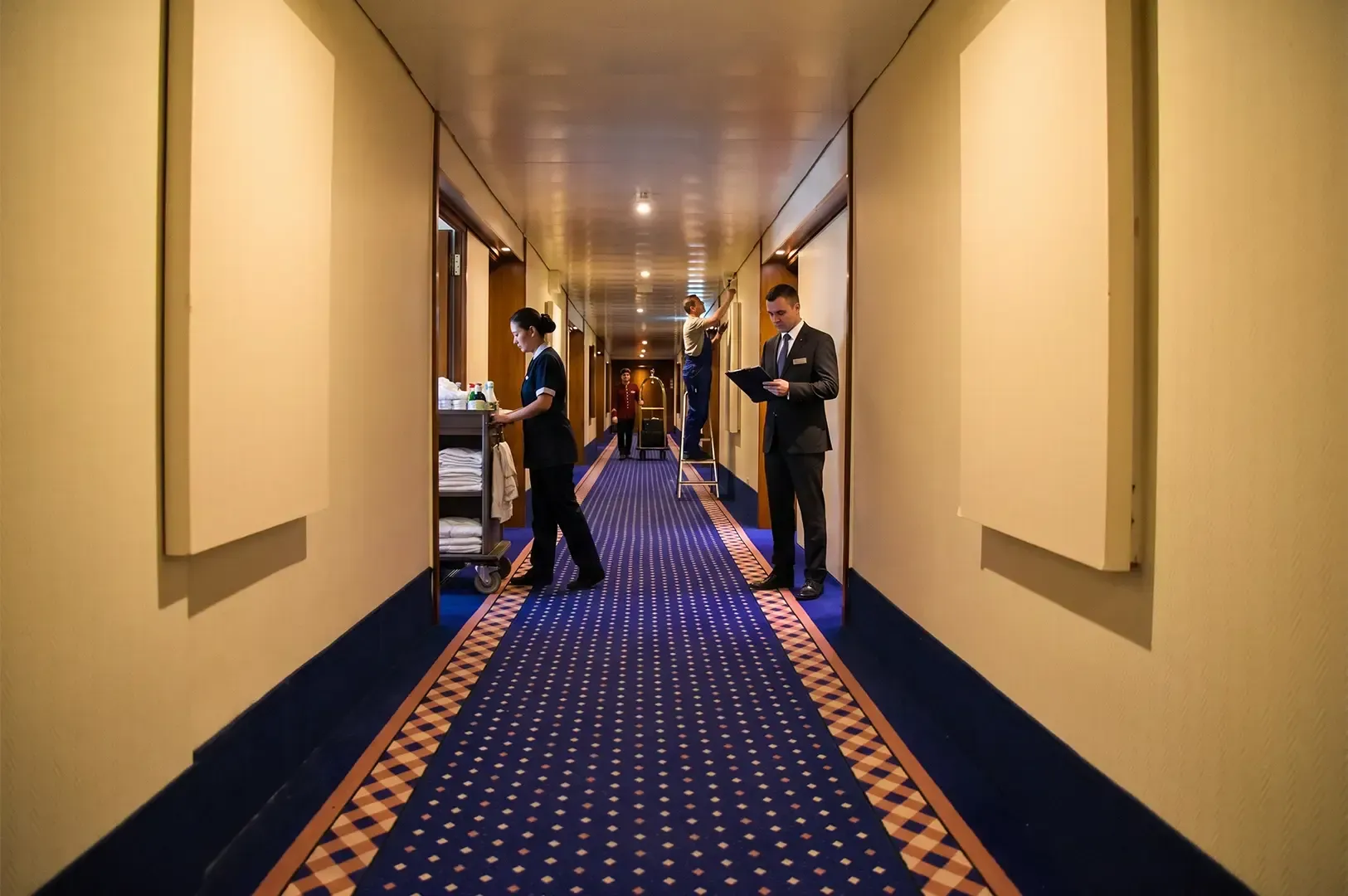 A long corridor with light yellow walls and blue and orange patterned floor, with hotel staff doing their duties all over the corridor.