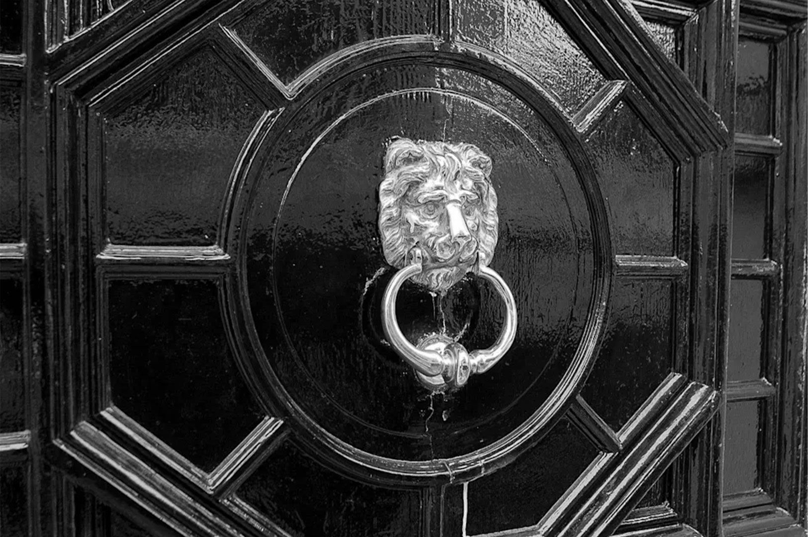 A close-up, black and white photograph of a glossy, paneled black door with a traditional silver lion head door knocker and ring.