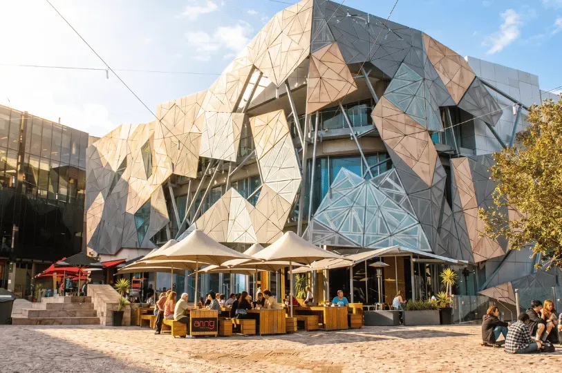The geometric, geometric-tiled facade of the ACMI building at Federation Square in Melbourne, with outdoor cafe seating under large umbrellas filled with people relaxing in the open plaza.