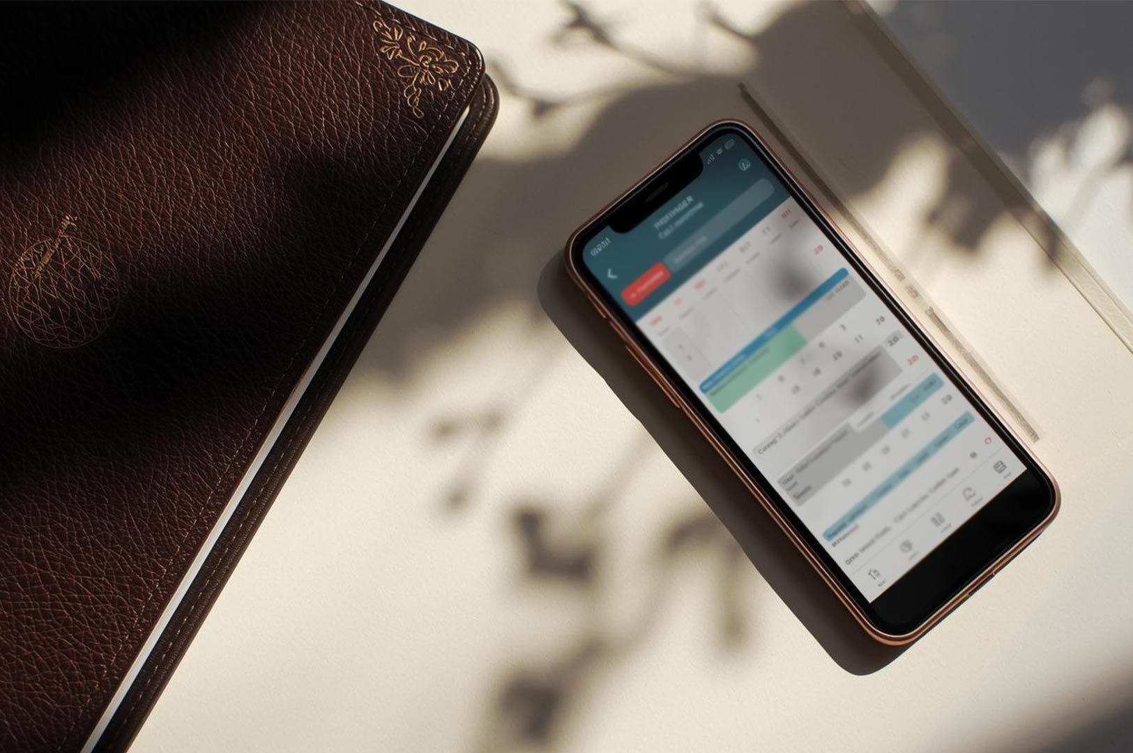 Overhead flat lay of a smartphone displaying a scheduling calendar app next to a brown leather notebook on a white desk, illuminated by dappled sunlight shadows.