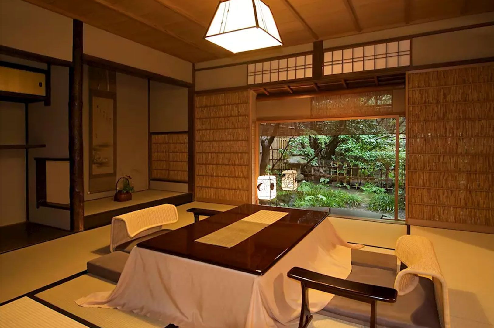 Interior of a traditional Japanese ryokan room featuring tatami mats, a cozy kotatsu heated table, and large sliding windows looking out onto a lush green garden.