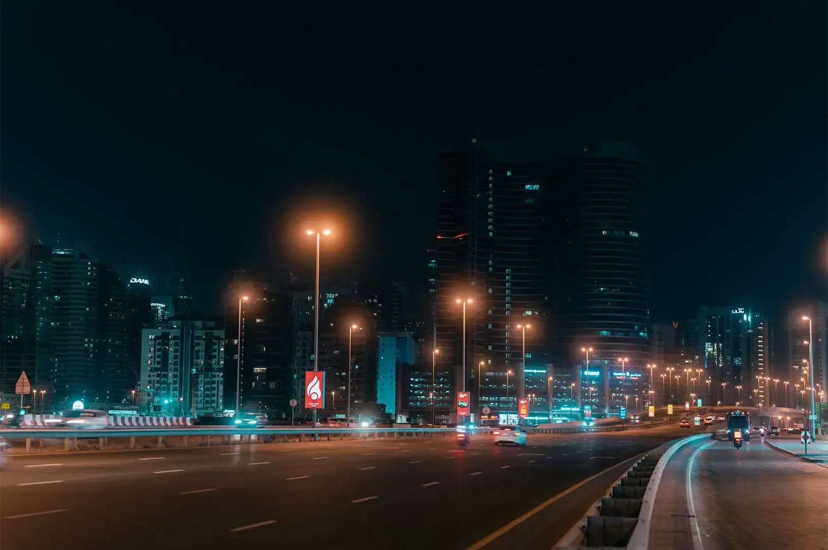 Dubai highways with a few cars and motorcycle driving along the road lit by warm street lights under a night sky with the city at the background.