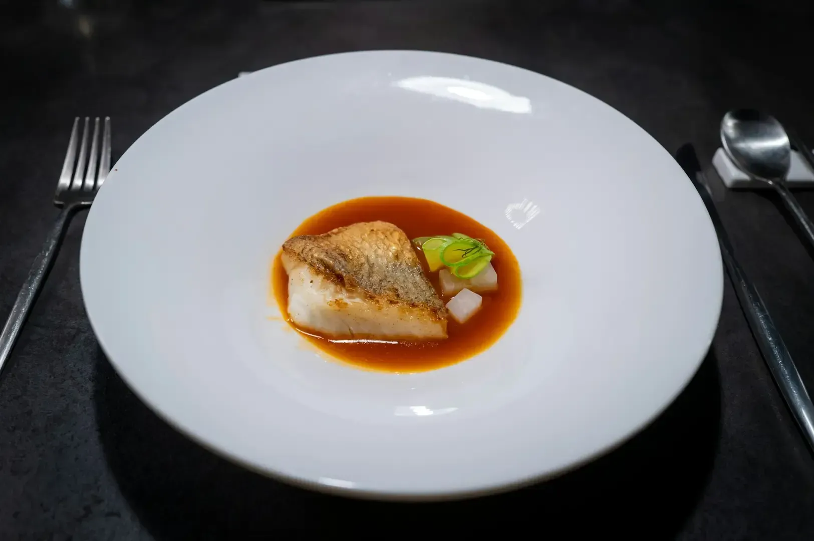 A simple fine dining dish on a white plate with utensils neatly placed on the sides on top of a black textured table.