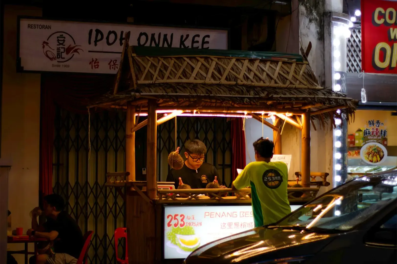 A hawker cart by a busy street with a wooden style build, a lone customer ordering from a staff behind.