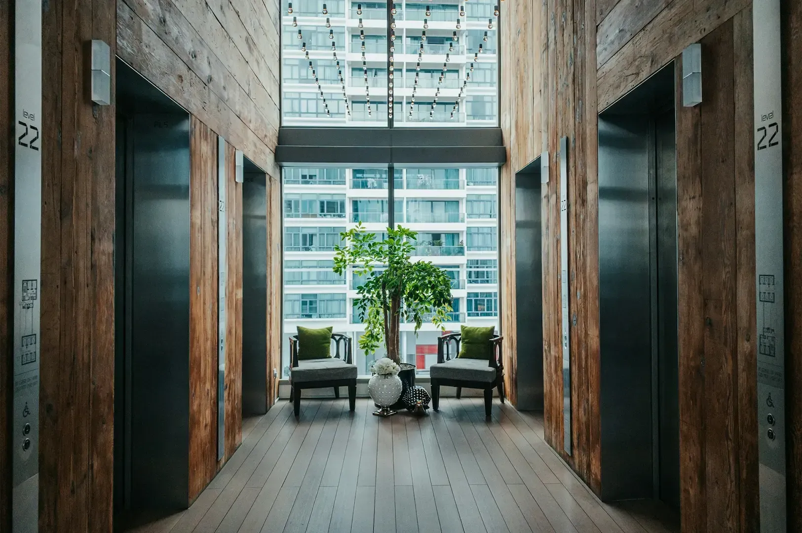 Wide shot view of four lifts in a room with wooden finish, decorated with chairs and plants with a window giving view to a high-rise building behind.