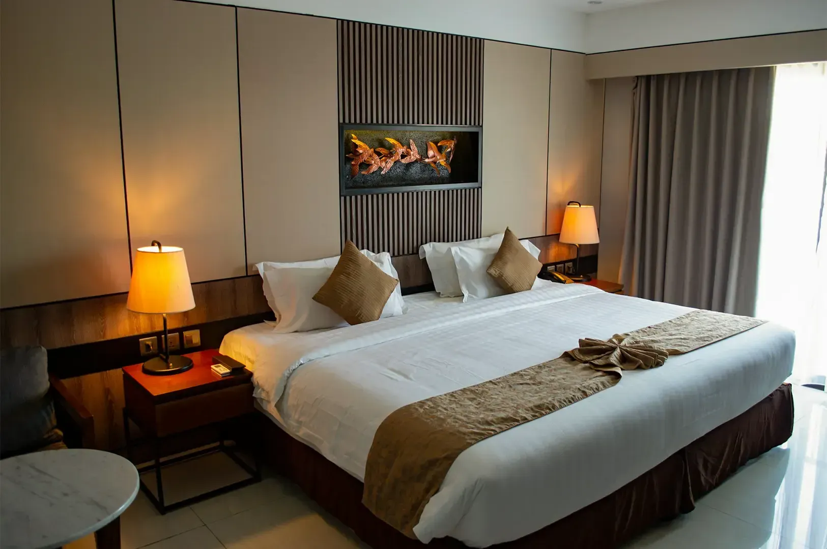 A hotel room interior with a bed with two lamps on either side, slightly parted curtains on the right letting in a soft sunlight.