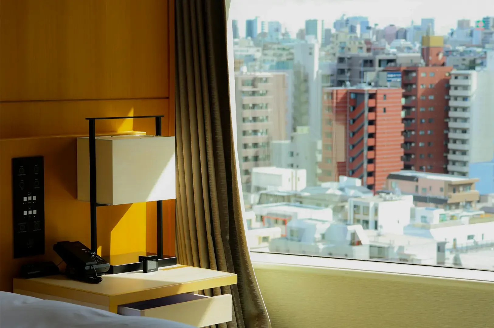 Hotel room interior focused on its bedside window with a city view outside in broad daylight.