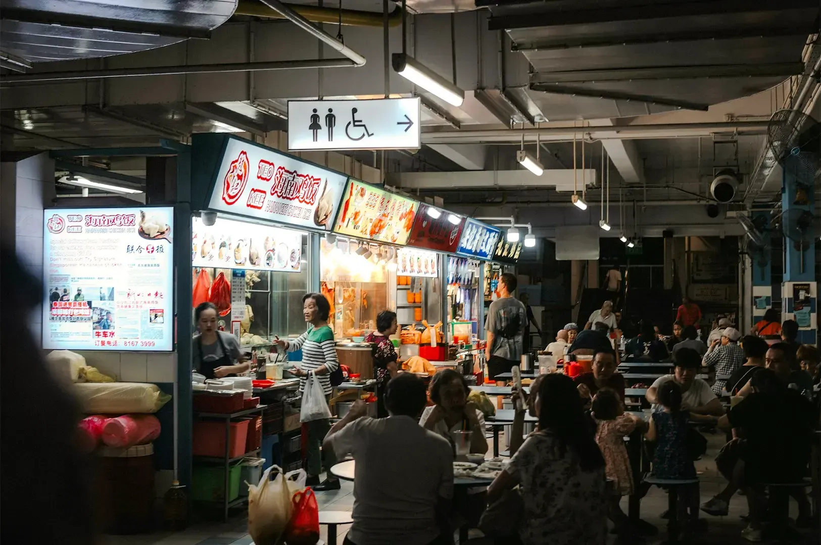 A cozy and dimly lit hawker place with stalls lined on the left with white and yellow lights reflecting off dining customers at the side.