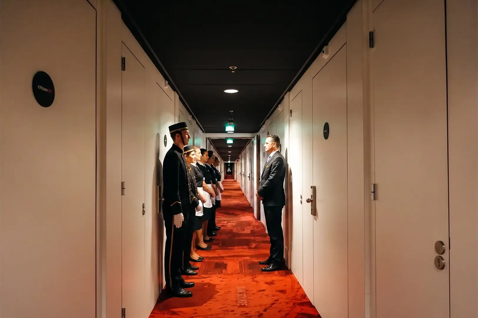A group of hotel staff lined up in a corridor with white walls and doors and orange tiled floor.