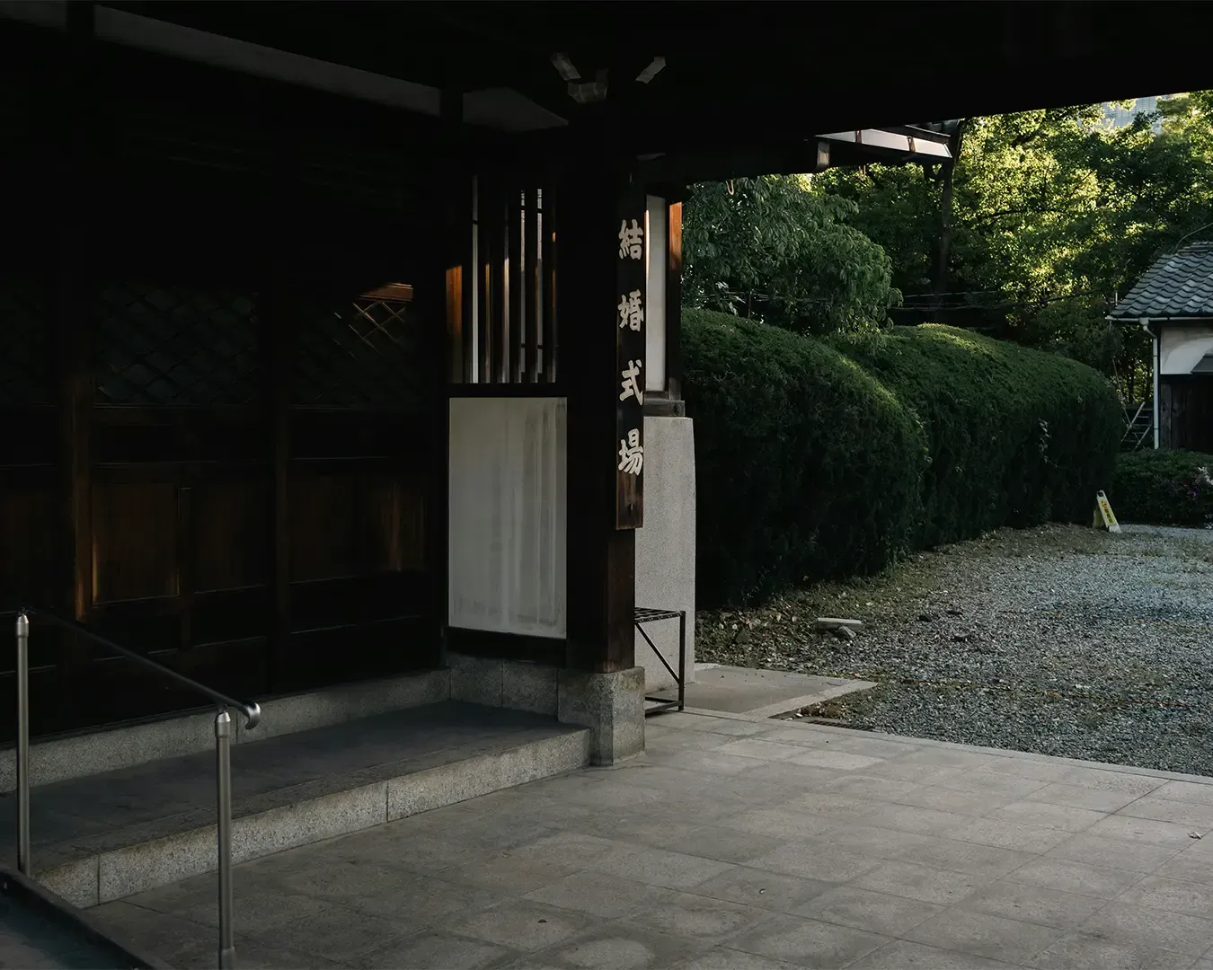 Entrance to a traditional Japanese building with wooden doors and a sign. A pathway lined with green bushes leads to a serene garden area.