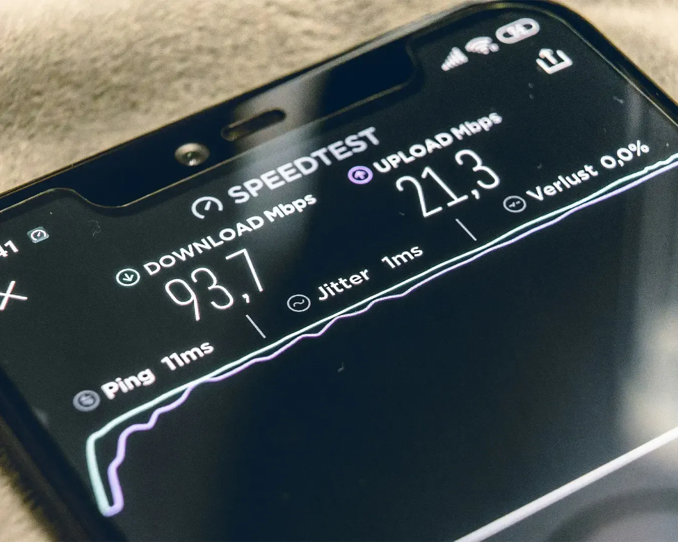 A smartphone held in hand displaying an internet speed test on the screen, with a speedometer-style graphic measuring connection performance.