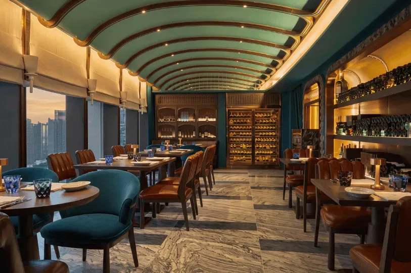 An upscale restaurant interior characterized by a distinctive vaulted teal ceiling with gold trim. The warm room features dark wood tables, a mix of teal and brown leather chairs, an illuminated glass wine cabinet, and large windows showcasing a city sunset.