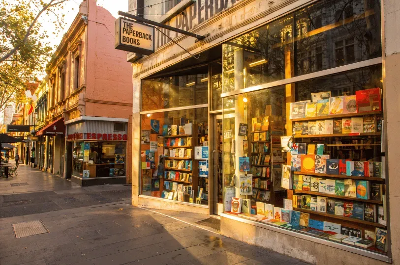 The street-level storefront of The Paperback Books on Lygon Street in Melbourne, showing glowing floor-to-ceiling glass windows filled with curated book displays during golden hour.