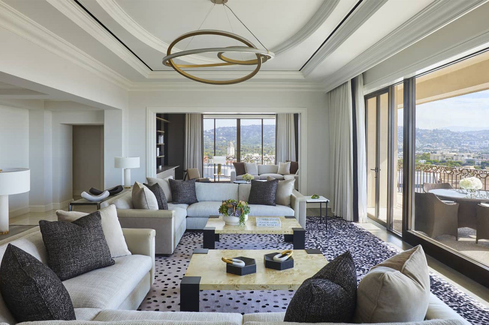 A spacious, modern luxury suite living room with light-colored modular sofas, a unique chandelier, and large glass doors leading to a balcony with a panoramic hill view.