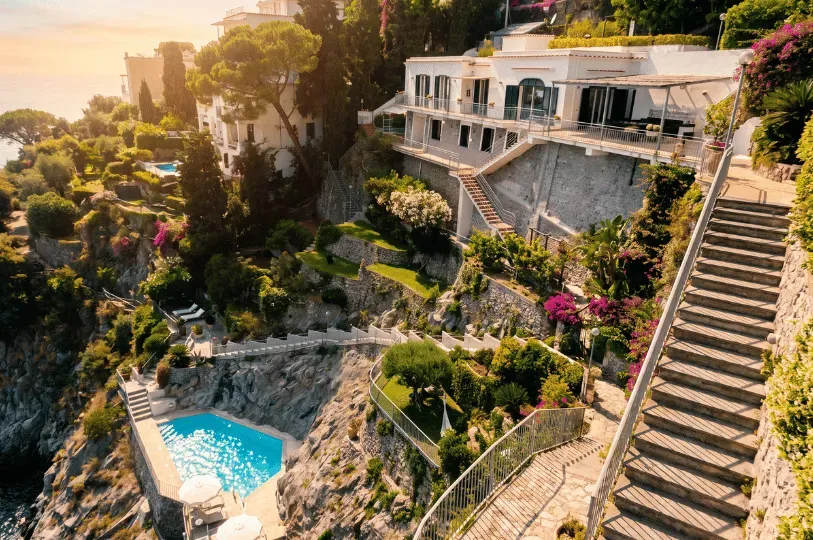 A bright white Mediterranean-style villa built into a steep, rocky cliffside overlooking the ocean. The property features extensive terraced gardens, long winding staircases, vibrant pink bougainvillea, and a private blue swimming pool on a lower level.