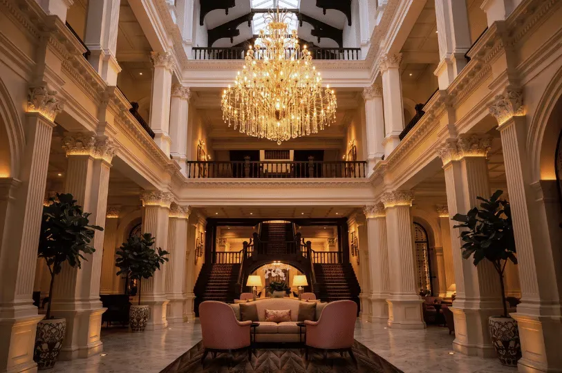 A spectacular multi-story hotel lobby with classic white pillars, a grand wooden staircase, and a massive crystal chandelier glowing with warm, luxurious ambient light.
