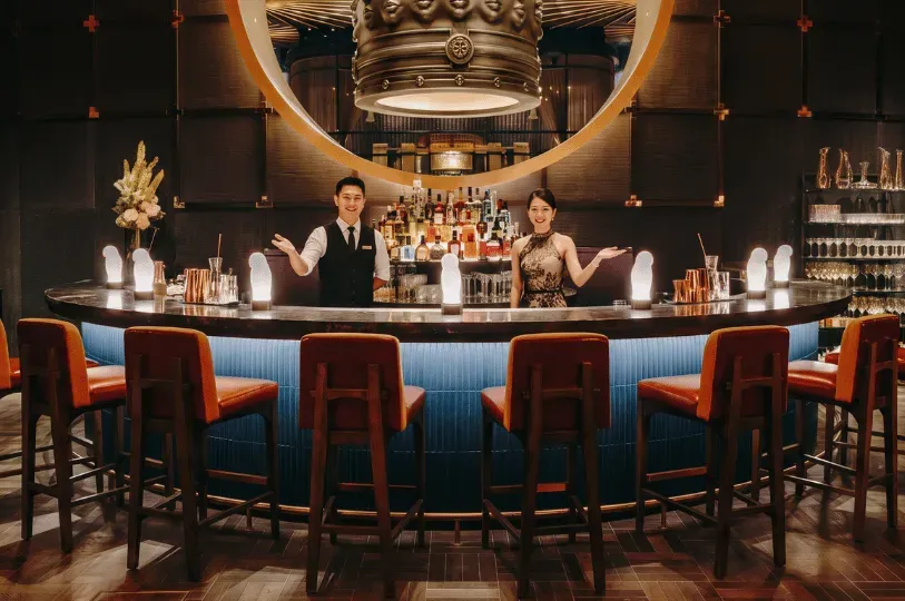 Two formally dressed bartenders smiling warmly and gesturing a welcome from behind an elegant, curved bar counter equipped with orange leather stools in a luxurious, dimly lit lounge.