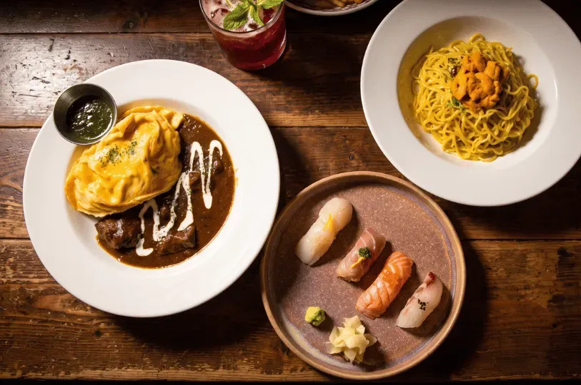An overhead shot of three distinct plates resting on a dark wooden table: a plate of omurice covered in a rich, dark brown demi-glace, a small ceramic plate featuring four pieces of delicate nigiri sushi, and a bowl of yellow noodles topped with bright orange sea urchin.