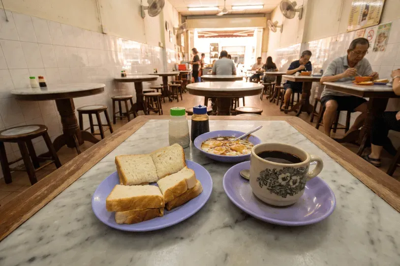 A first-person view of kaya toast, soft-boiled eggs, and black coffee on a marble kopitiam table. Local patrons dine in the softly blurred background of the old-school coffee shop.