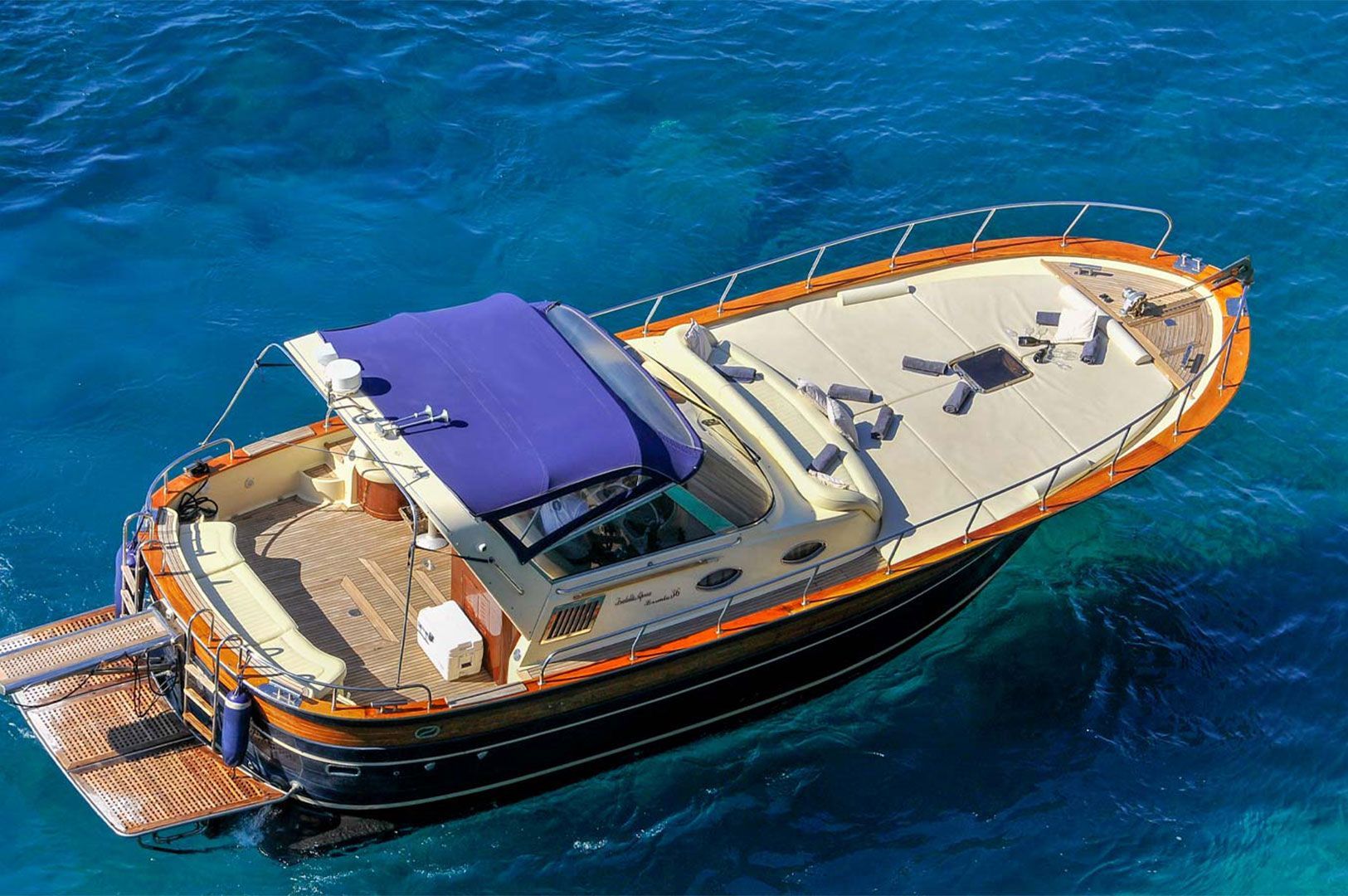 An aerial view of a classic wooden motorboat with a blue canopy sitting on the clear, turquoise water of the Mediterranean Sea.