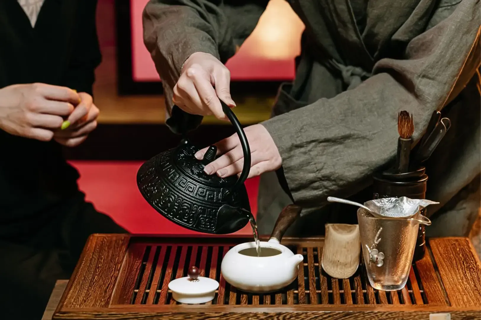 A medium shot of two people in traditional Japanese clothing pouring tea using a black tea pot in a white dinnerware on top of a wooden tray.