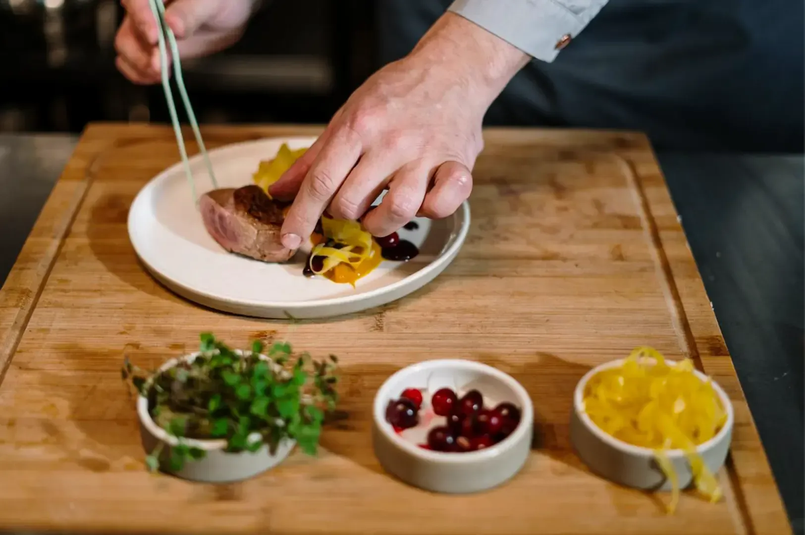 A chef preparing a dish with fresh herbs, berries, and cooked meat on a wooden tray.