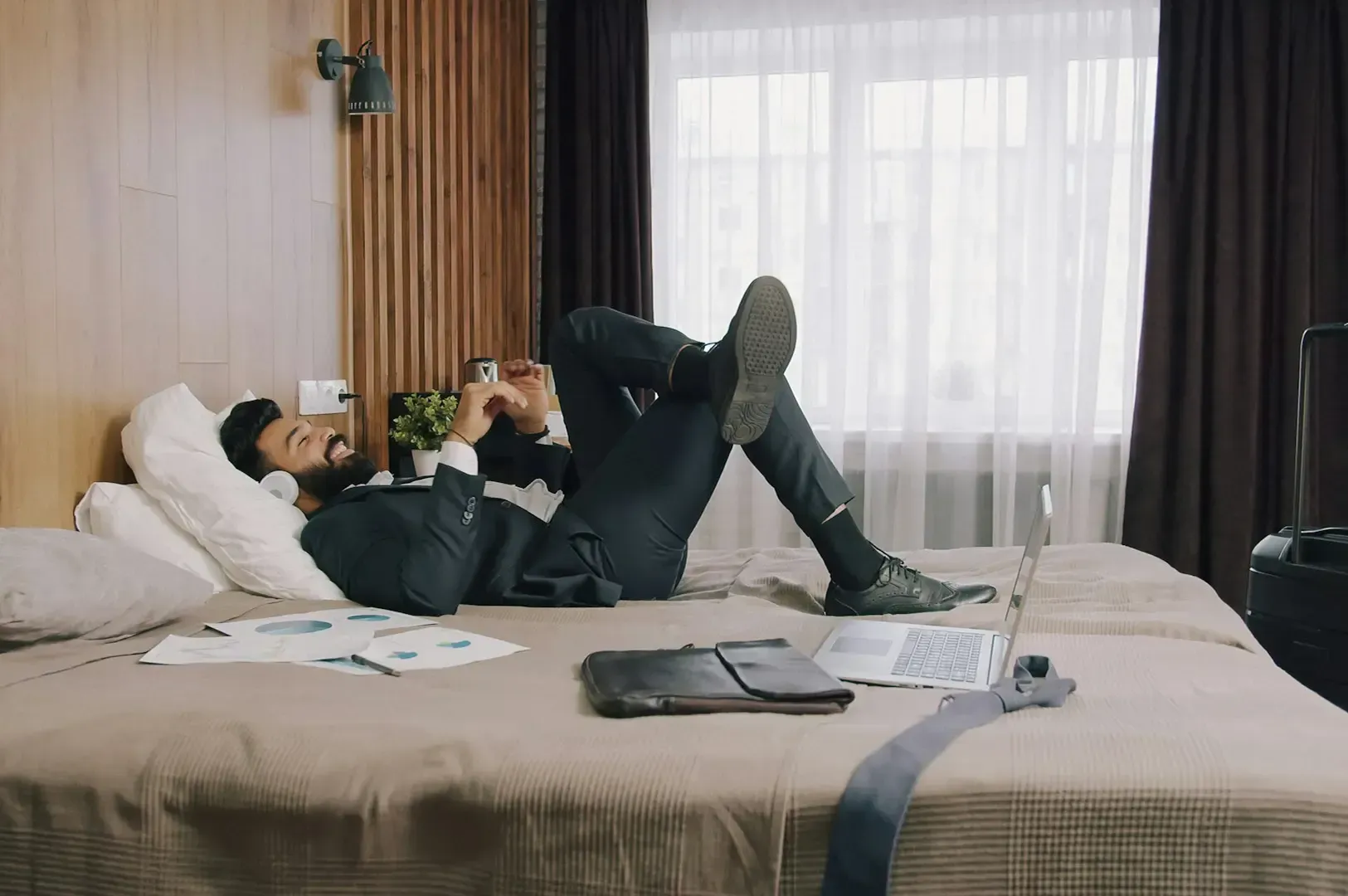 A hotel guest wearing a business attire laying relaxed on a bed with headphones and a laptop, papers, and a leather bag placed beside him, a parted curtain in front of a widow at the back.