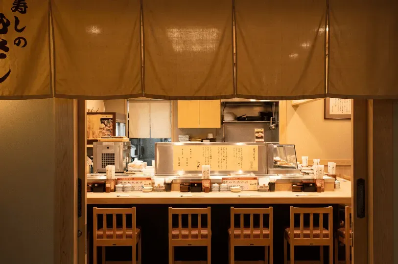 A warm, inviting Japanese sushi counter viewed through a traditional split fabric curtain. The minimalist light wood counter is neatly set with placemats and chopsticks, lined with simple wooden chairs, with a brightly lit preparation area visible in the background.
