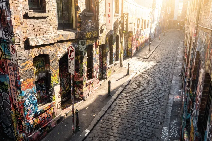 An elevated perspective looking down a cobbled Melbourne laneway, featuring brick walls covered in vibrant street art and graffiti under warm, streaming sunlight.