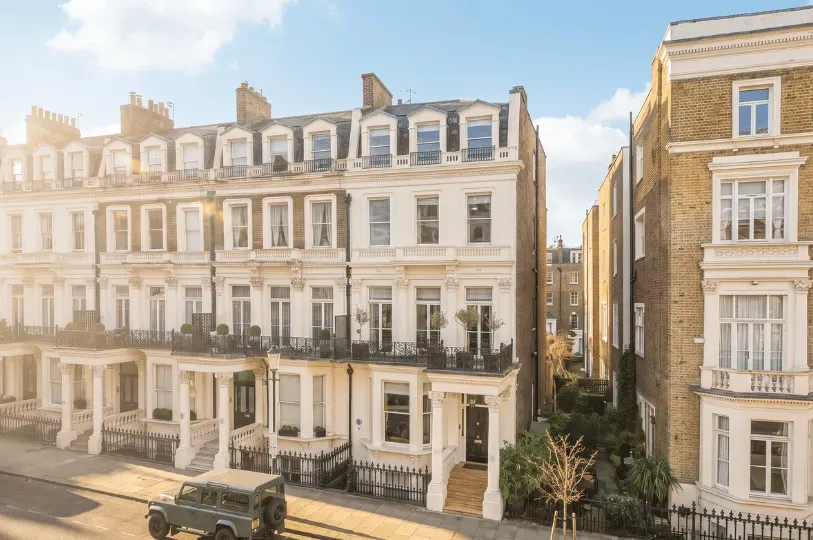 A street view of classic, elegant townhouses with cream-colored stucco facades, grand entrance columns, and black wrought-iron balconies. A green off-road vehicle is parked on the street in front of the multi-story residences on a sunny day.