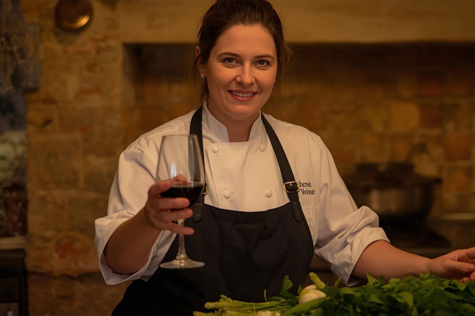 A smiling female chef in a white uniform and dark apron holds up a glass of red wine in a rustic, brick-walled kitchen setting, surrounded by fresh green vegetables.