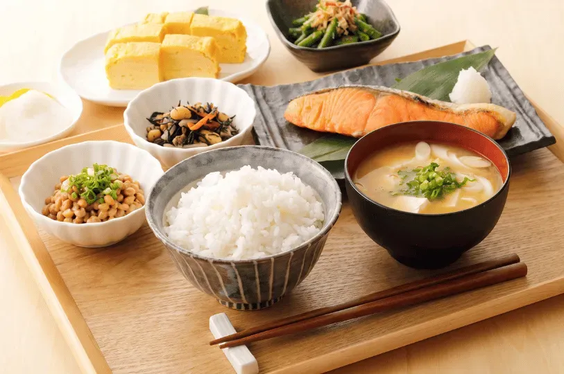 A traditional Japanese morning set meal thoughtfully arranged on a wooden serving tray. The spread includes white rice, miso soup, grilled salmon, tamagoyaki, and natto.