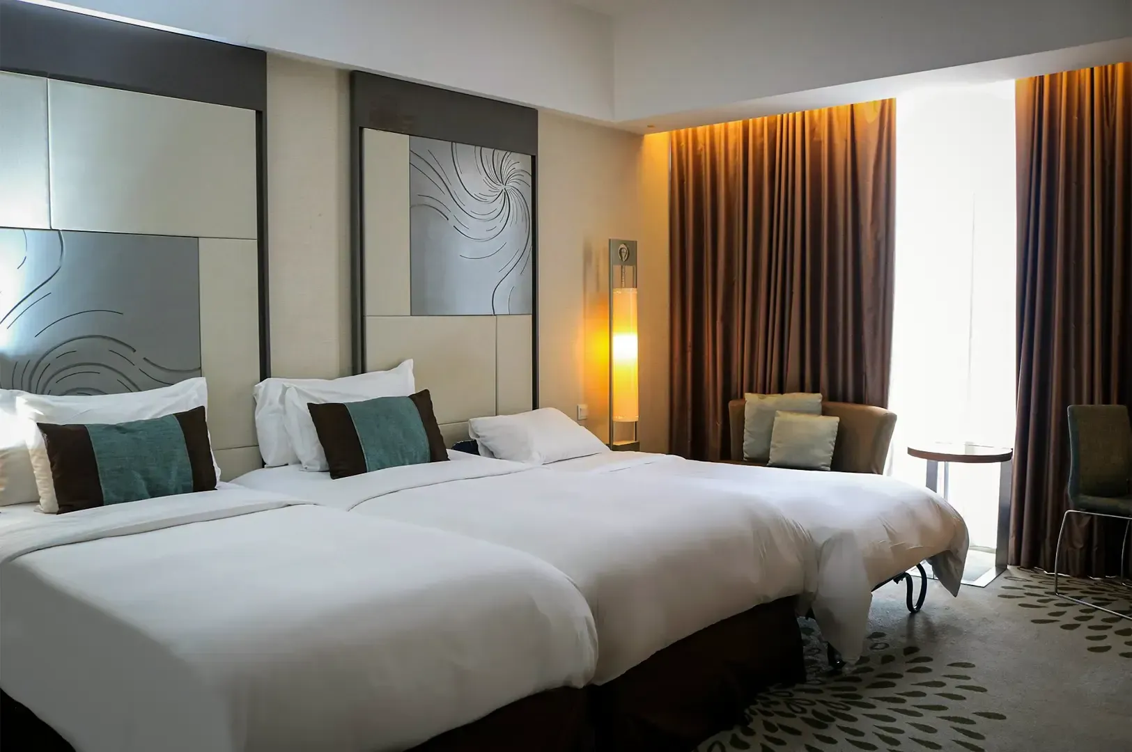 Wide angle shot of a hotel room with two white beds and curtains parted at the middle, with walls and floor decorated by modern designs and patterns.