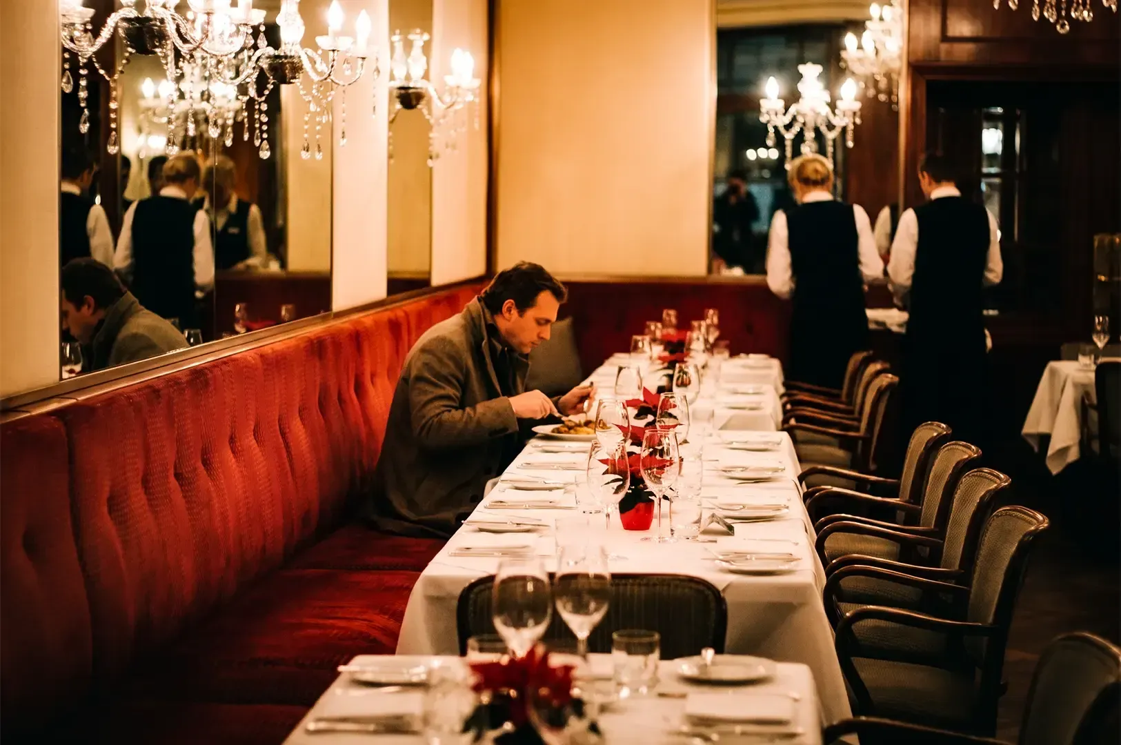 A lone customer sitting on a red cushioned seat eating on a fine dining table decorated with flowers on a vase and white cloth with waiters working in the background.