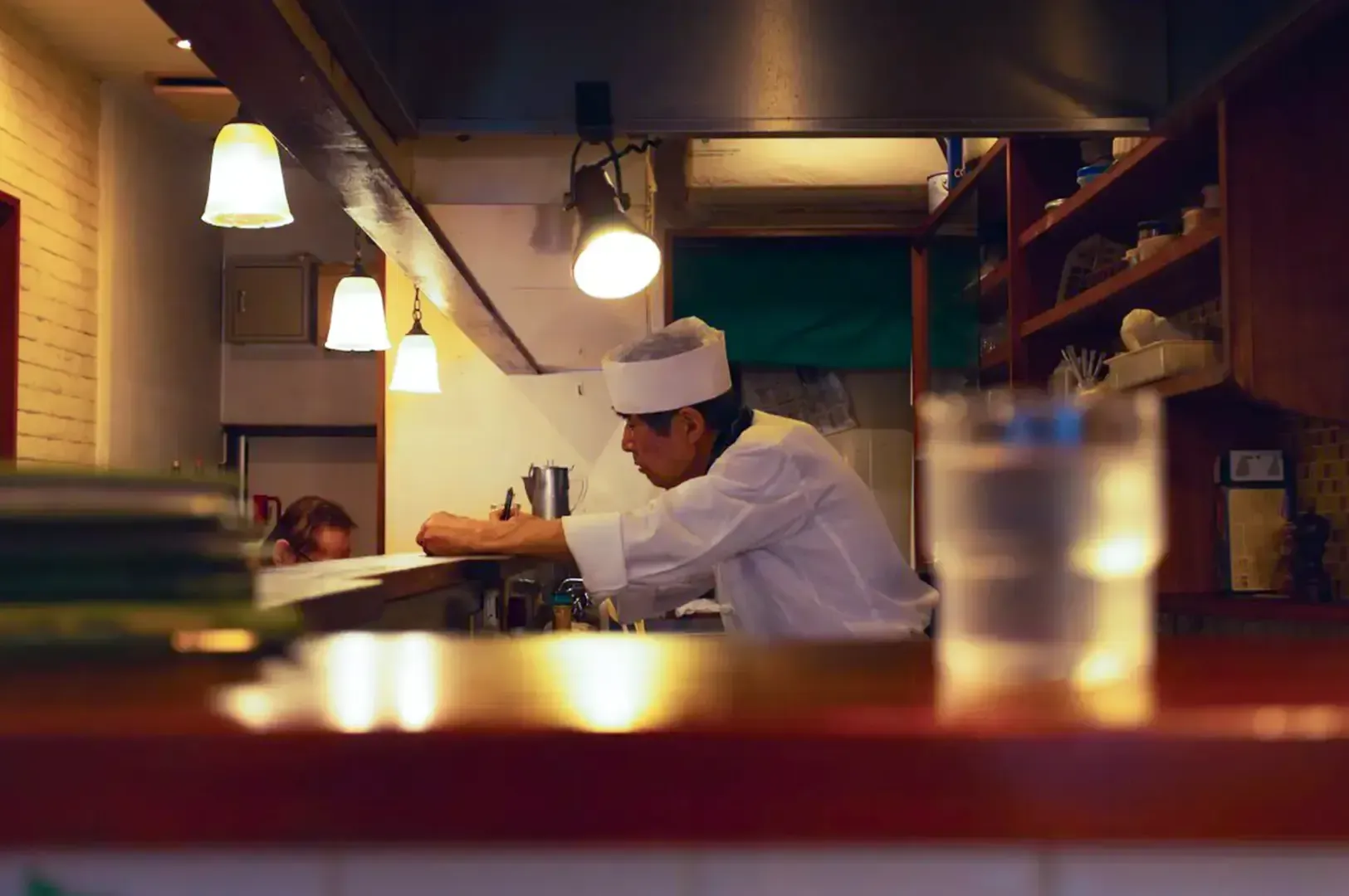 A lone chef in white uniform writing by his counter lit by warm yellow lights hanging above.