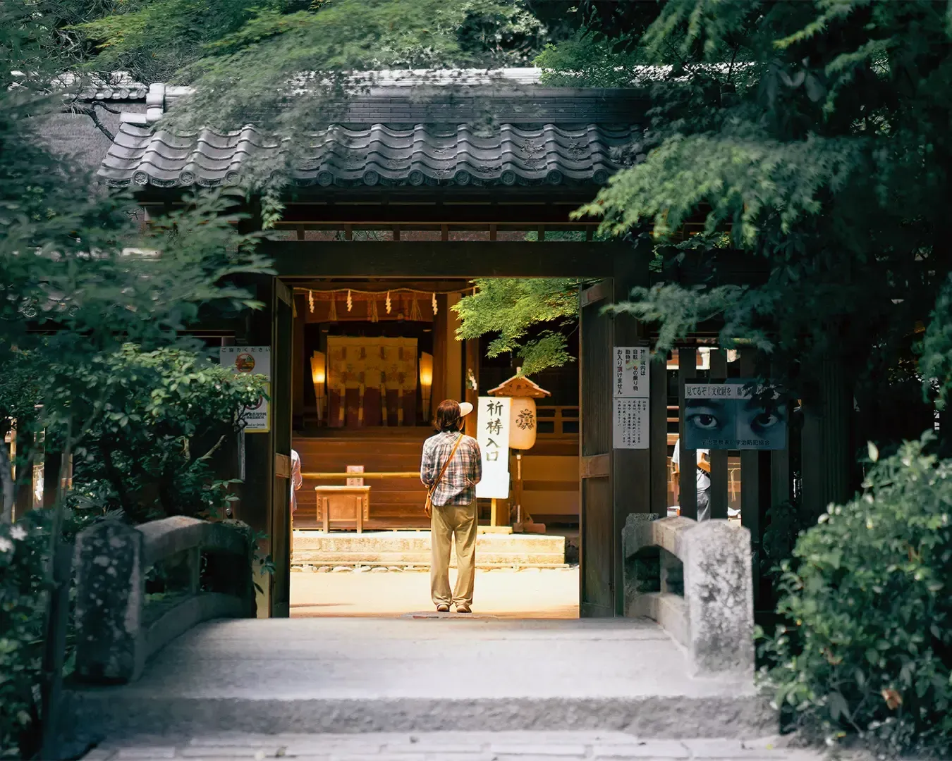 A person stands contemplatively at a wooden Japanese temple entrance, surrounded by lush greenery, conveying a peaceful, serene atmosphere.