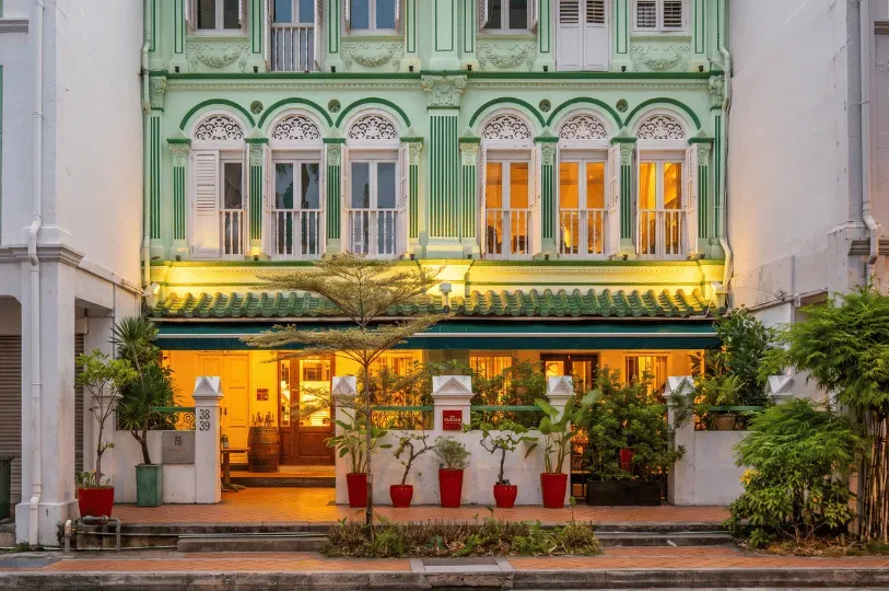 The illuminated exterior of a restored heritage shophouse at dusk. The building showcases an ornate pastel green and white upper facade with arched shuttered windows, contrasting with the warm, inviting glow of the ground-floor restaurant framed by potted plants and a tiled walkway.