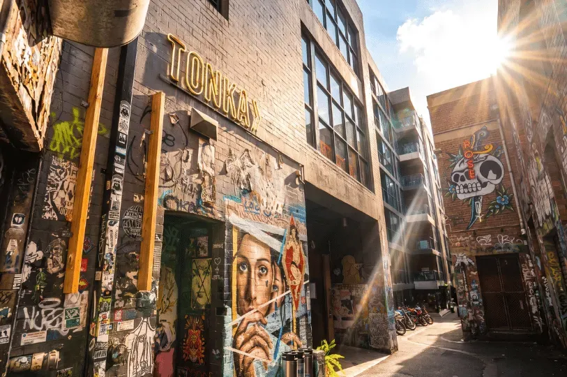 A sunlit view of Duckboard Place in Melbourne, featuring a wall with graffiti art, a yellow neon sign for Tonka restaurant, and a skull-and-rose mural illuminated by a bright sun flare.