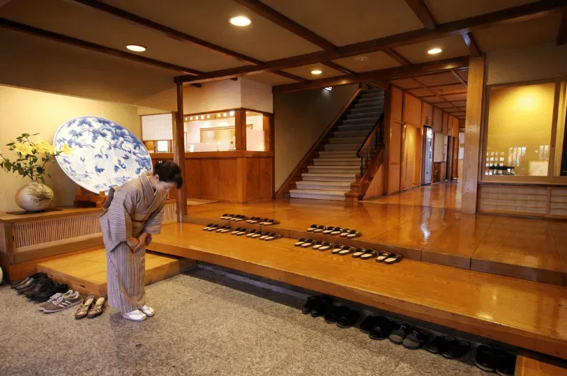 The traditional entryway of a Japanese ryokan or residence with a polished wooden floor. A woman wearing a striped kimono bows deeply in greeting. Neatly arranged slippers sit on the raised wooden step, while outdoor shoes remain on the lower stone floor near a large decorative ceramic plate.