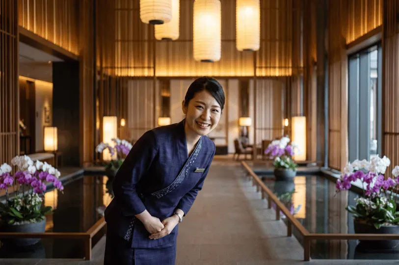 A smiling hotel staff member offering a warm, welcoming bow in a serene, softly lit lobby decorated with reflective water features and vibrant purple orchids.