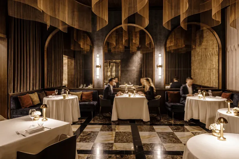 An upscale, dimly lit fine-dining room featuring plush blue velvet booths, crisp white tablecloths, glowing table lamps, and mirrored walls reflecting a couple enjoying a quiet meal.
