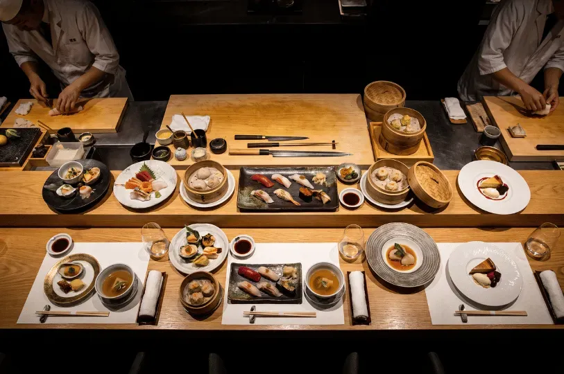 A symmetrical overhead view of an upscale chef's counter. At the top edge, the hands of two chefs are visible preparing ingredients on cutting boards. The center of the counter is lined with shared platters of sushi and bamboo steamer baskets, while perfectly arranged individual place settings with small tasting dishes sit waiting for diners along the bottom edge.