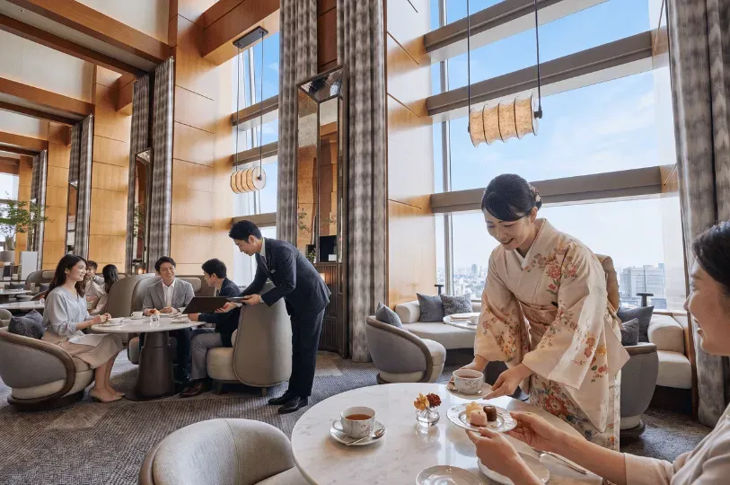 A bright, high-ceilinged luxury hotel lounge with expansive city views, where a smiling server in a traditional floral kimono serves tea and pastries to a guest, while a suited staff member assists another table in the background.