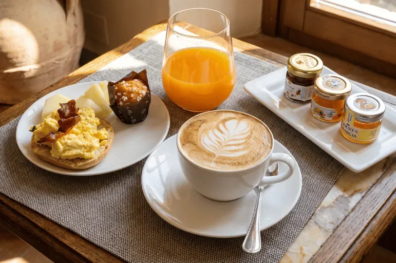 A bright continental breakfast set by a sunlit window with a cappuccino and orange juice. The meal features scrambled eggs and bacon on a muffin alongside fruit preserves.