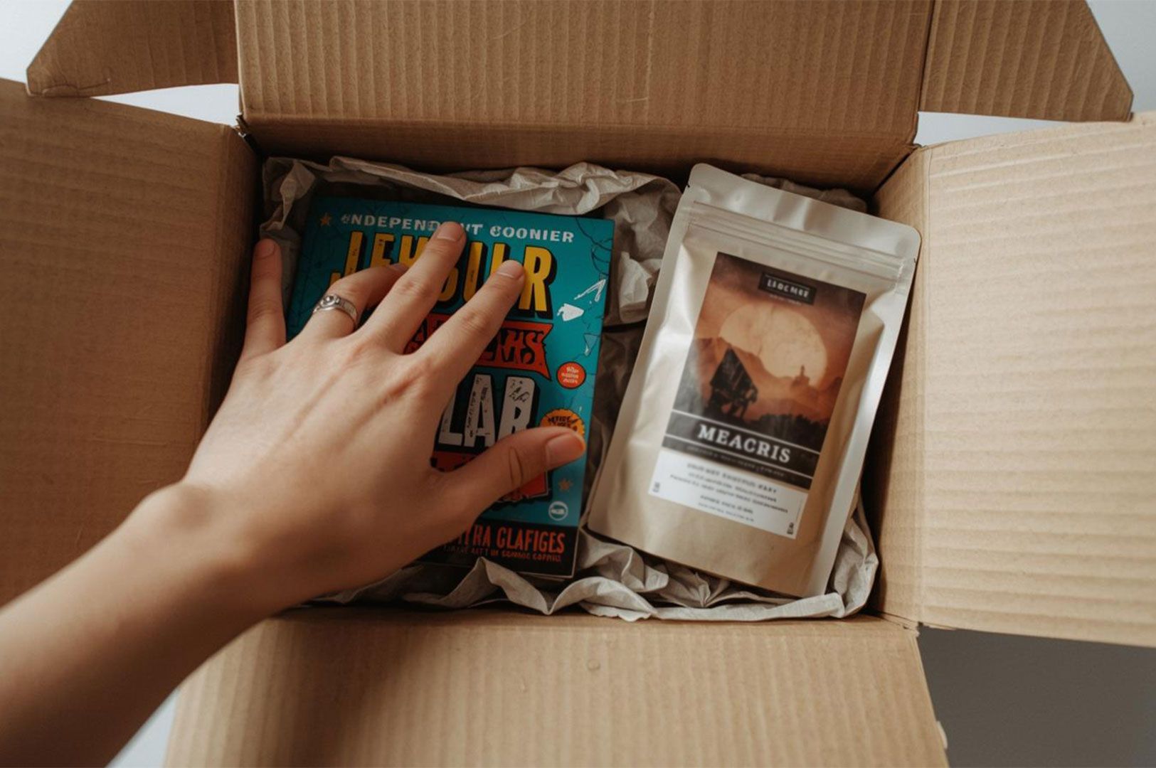 A top-down perspective of a hand reaching into an open cardboard shipping box containing a colorful book and a pouch of gourmet coffee.