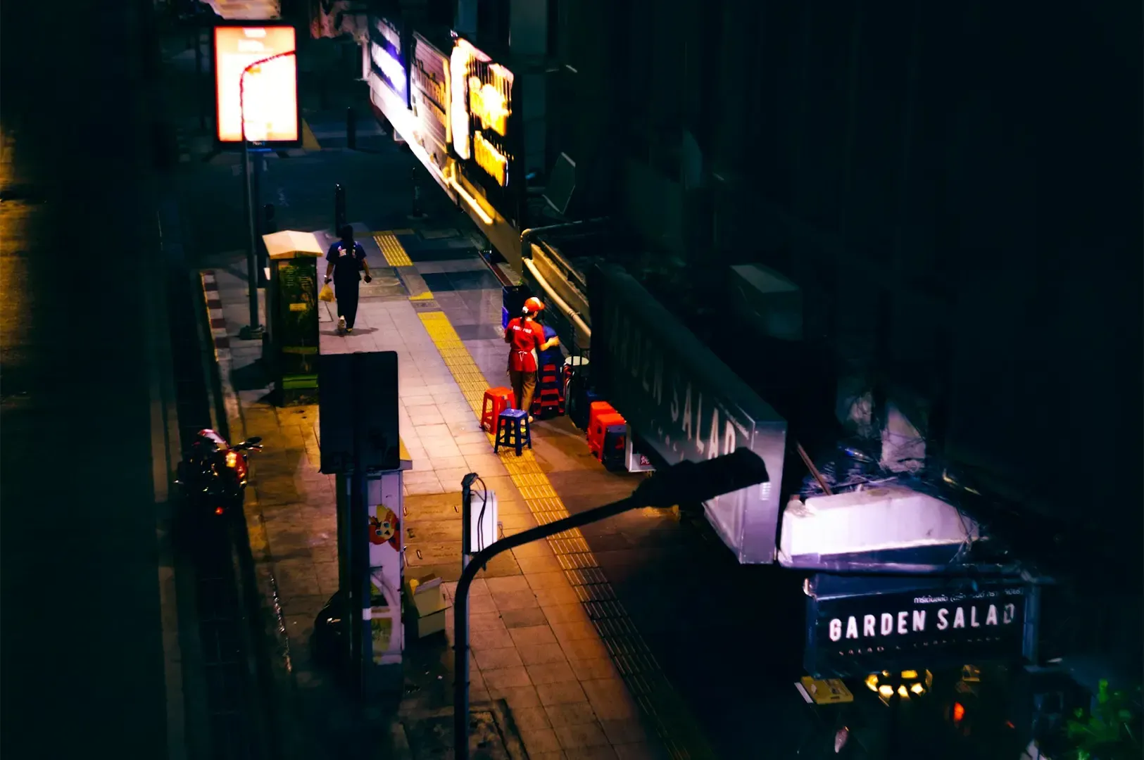 A dimly lit street in Bangkok with a few street lights and shop lights lighting the sidewalk.