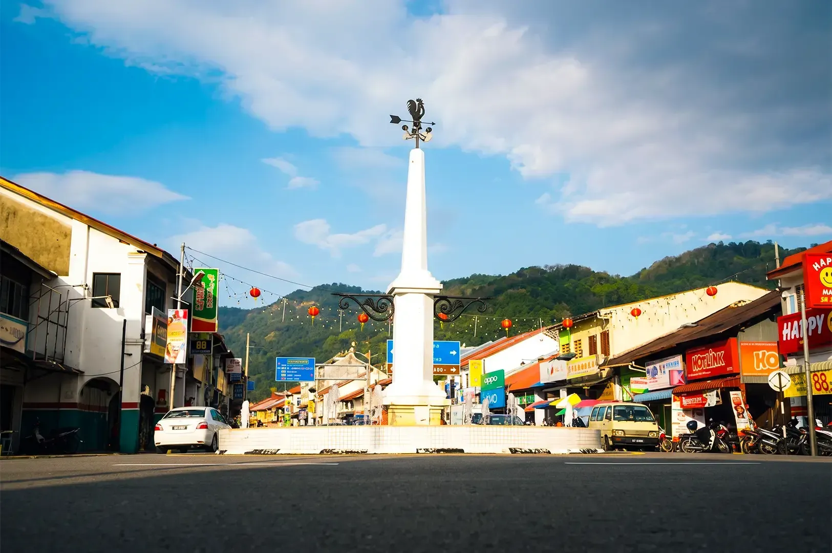 Balik Pulau with a central white monument focused on the center and various shophouses lined either side of the street with green mountains visible in the background.