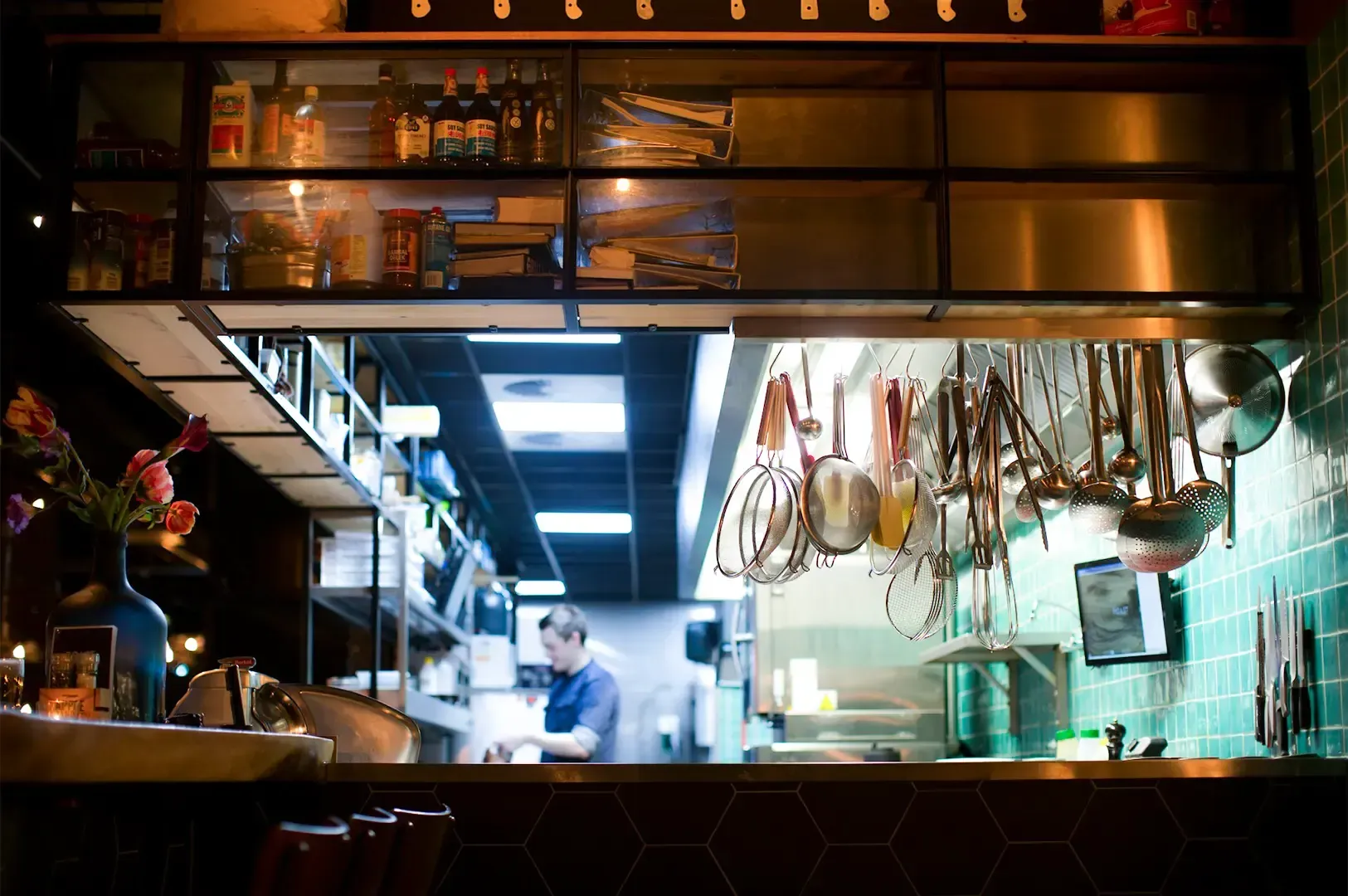 A wide angle view of a dim and warmly lit counter with bottles placed on shelves above and a chef working behind.